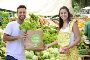 Small business owner at an open street market, selling organic fruits and vegetables to a woman carrying a shopping paper bag with a 100% organic certified label.